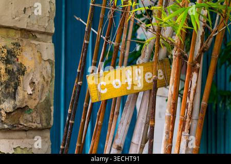A weathered yellow wooden sign with "coffee" written in white paint, hanging from vertical bamboo poles against a blue and textured stone wall backgro Stock Photo