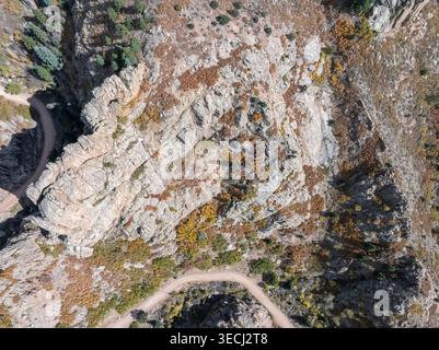 Aerial view of a steep rocky cliff face with colorful autumn foliage and a winding dirt road through the mountain terrain. Stock Photo