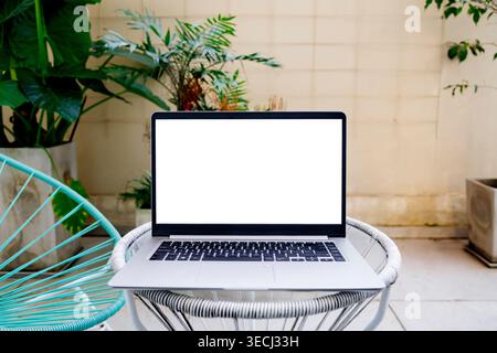 Laptop for Mockup with Blank White Screen on Outdoor Table in Modern Garden Patio with Tropical Plants and Acapulco Chair Stock Photo