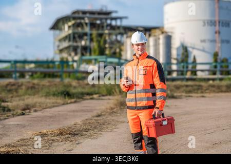 Mid-adult Caucasian male engineers in orange safety gear inspect an industrial plant for energy sector logistics, maintenance, and professional workpl Stock Photo
