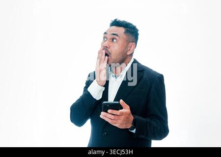 An Indonesian man in a black suit holding a smartphone with a shocked expression, hand over mouth and eyes wide, isolated on a white background. Stock Photo