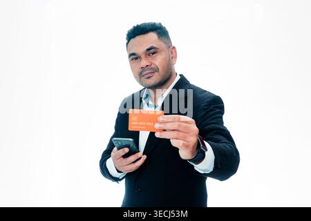 A professional Indonesian man in a black suit holding an orange credit card and a smartphone, isolated on a white background. Stock Photo