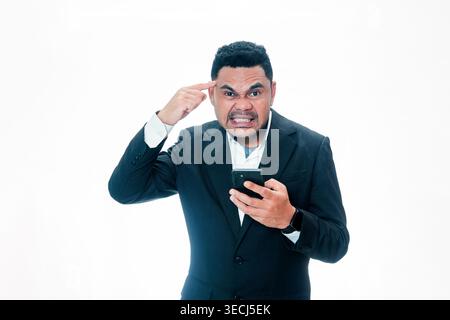 A frustrated Indonesian man in a black suit holding a smartphone and pointing at his head with a stressed facial expression on a white background. Stock Photo