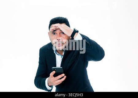 An Indonesian man in a black suit holding a smartphone and shielding his eyes to look far away, representing vision and searching for opportunities. Stock Photo