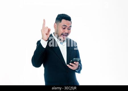An Indonesian man in a black suit looking at his smartphone with an excited, open-mouthed expression and pointing his finger up, isolated on a white b Stock Photo