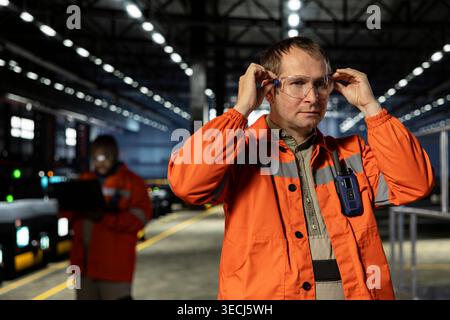 Male operator putting safety glasses and oversees assembly tools in factory, surrounded by steel machinery for welding within warehouse. Workforce involvement and heavy manufacturing tasks. Stock Photo