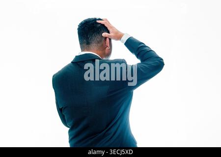 Back view of an Indonesian man in a black suit holding his head thoughtfully, expressing confusion or decision making on a white background. Stock Photo