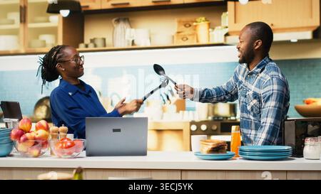 Black young couple starting a duel with kitchen utensils for the game, playful fight with fake swords during the goofy war. Childish silly adversaries having fun together in the morning. Stock Photo