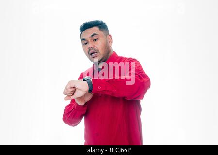 An Indonesian man in a red shirt looking at his smartwatch with a shocked and worried expression on a white background. Stock Photo