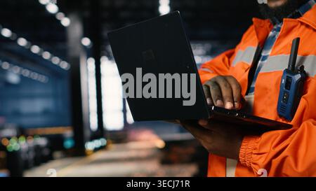 African american supervisor directing production tasks using laptop in a safety vest, surrounded by heavy machinery, tools and construction defining modern industrial manufacture. Stock Photo