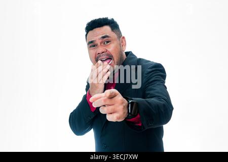 Portrait of a joyful man in a black suit and red shirt laughing heartily while pointing his finger directly at the camera, with one hand covering his Stock Photo