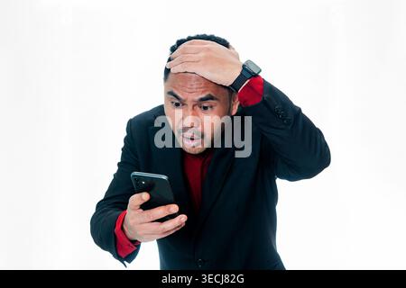 Portrait of a man in a black suit looking at his mobile phone with a stunned and disbelieving expression, holding his hand to his forehead, isolated o Stock Photo