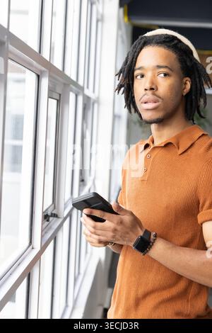 African American man standing by windows in hall holding device while wearing rust polo, copy space Stock Photo