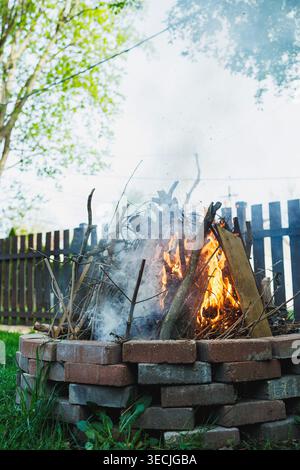 A campfire burns brightly in the backyard. Smoke rises from the fire as sticks and wood crackle. The sunlight shines on the green grass and wooden fen Stock Photo