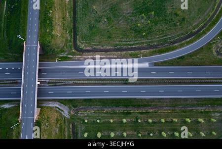 Aerial view of a multi-lane highway interchange featuring a bridge overpass, lush green fields, and organized rows of trees. Stock Photo