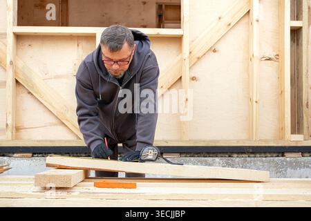 Carpenter man working with timber at a construction site. Frame house construction, DIY eco wooden home concept. Skilled worker measuring beam outdoor Stock Photo