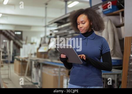 Female African American manager taking inventory in a metal factory using a tablet for modern digital supply chain management in heavy industry. Stock Photo