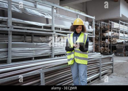 Female worker in a metal products factory taking inventory of goods and checking the stocks in an open warehouse using a digital tablet. Stock Photo