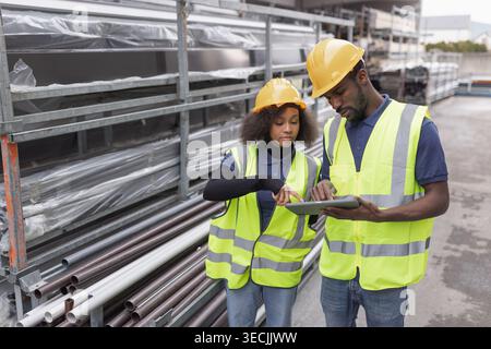 Two workers wearing protective gear, yellow hard hats and safety vests taking inventory in an outdoor warehouse using a digital tablet. Stock Photo