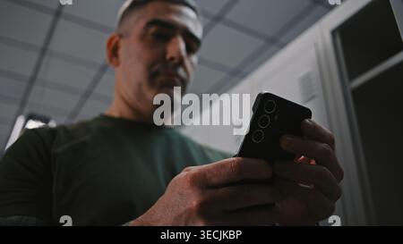 Soldier Stands in Military Locker Room, Using Smartphone to Send Updates and Stay Connected Preparing Between Training Sessions. Integration of Modern Communication Technologies Into Military Routine. Stock Photo