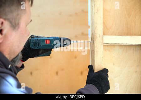 Professional unrecognizable constructor using electric screwdriver to fix wooden plank. Work in progress,  frame house under construction. DIY eco woo Stock Photo