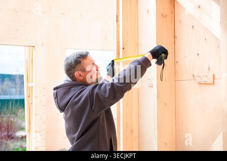 Professional constructor using using ruler at frame house under construction. Work in progress. DIY eco wooden home concept. Skilled builder man with Stock Photo
