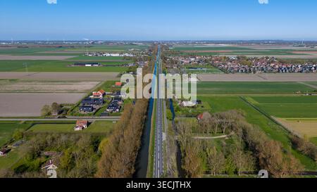 Aerial view of a long straight road and canal running through flat agricultural fields and residential areas under a clear blue sky in the Netherlands Stock Photo