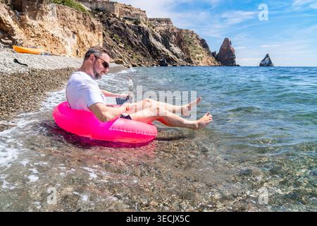 Freelancer laptop beach man using computer while sitting in a pink inflatable ring at the edge of the sea by rocky cliffs Stock Photo