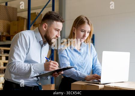 A professional team of logistics managers, a man and a woman, working together in a warehouse. They are checking inventory records using a laptop and a clipboard for efficient stock control. Stock Photo