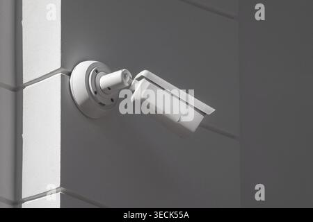 White security camera mounted on a wall corner. Cylindrical lens faces outward, recording surroundings. Surrounding tiles form sharp, geometric pattern. Soft light highlights camera's clean Stock Photo