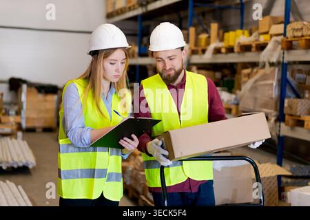 Concentrated warehouse workers examining open cardboard box while checking inventory list on clipboard. Systematic order verification and accuracy control Stock Photo