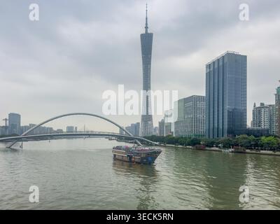 Guangzhou, China - March, 20, 2026: Grey sky looms above modern skyline. River flows gently beneath arched bridge. Tall tower stands near glassy skyscrapers. Boat drifts calmly on water's surface. Urban landscape blends nature and steel Stock Photo