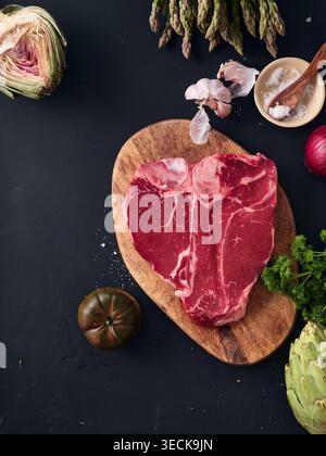 A raw t-bone steak is placed on a wooden cutting board. Fresh vegetables and herbs surround the steak, showing garlic and asparagus in a kitchen setti Stock Photo