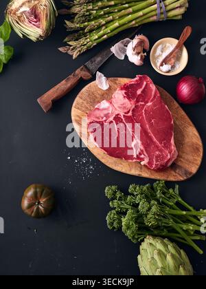 A raw t-bone steak is on a wooden cutting board. Surrounding it are fresh vegetables, herbs, and a knife. The setting is a kitchen with a dark surface Stock Photo