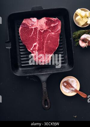 A raw t-bone steak sits on a black grill pan. Next to it are garlic cloves, butter, and a bowl of salt. This scene shows preparation for a meal. Stock Photo