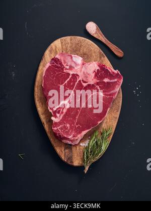 A raw t-bone steak sits on a wooden cutting board. Fresh herbs and salt are placed next to the steak. The setting is simple and clean, showing the raw Stock Photo