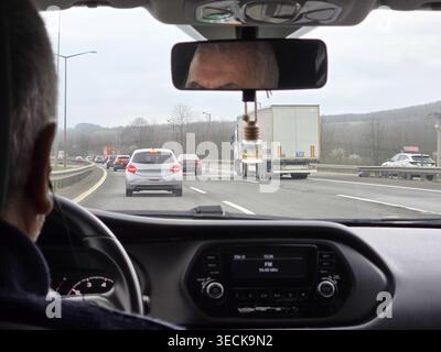 View from inside a car with driver silhouette and dashboard, looking through the windshield at heavy highway traffic with trucks and cars during a clo Stock Photo
