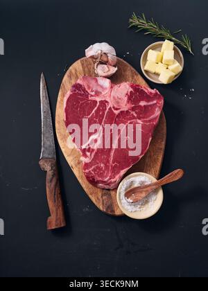 Raw t-bone steak sits on a wooden cutting board. A knife rests nearby, and bowls with butter, garlic, and herbs are arranged around it. The setting sh Stock Photo
