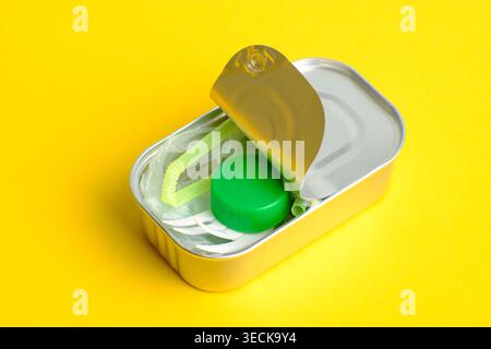 Open metal can packed with plastic trash like bottle cap, straw and bag on bright yellow background, visual metaphor for microplastic pollution, conta Stock Photo