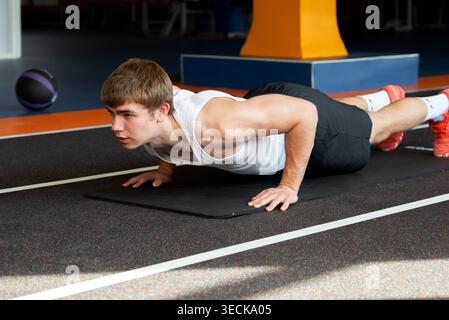 Young athlete doing push-ups on a mat in a gym, focusing on strength and endurance. Wearing a white tank top and black shorts, he embodies fitness and Stock Photo