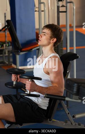 A young sportsman in a white tank top and black shorts lifts dumbbells in a modern gym. His focused expression highlights dedication and strength trai Stock Photo
