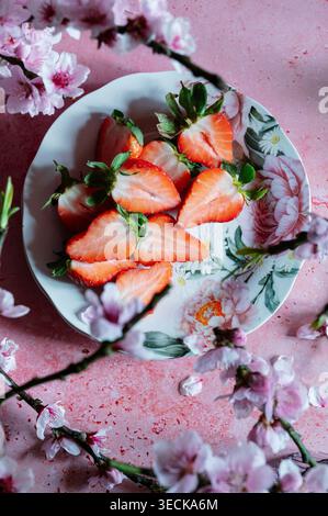 Overhead view of fresh strawberry halves arranged on a floral plate, styled with soft pink spring blossom and petals on a pastel background, creating Stock Photo