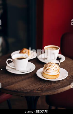 Warm cafe scene with two white coffee cups on saucers and a sweet pastry with toasted meringue on a wooden table, set against a soft red and dark back Stock Photo