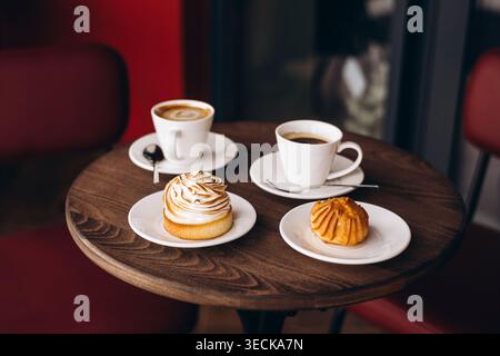 Warm cafe scene with two white coffee cups and saucers on a wooden table, paired with a lemon tart and a small pastry. Soft light and red chairs in ba Stock Photo