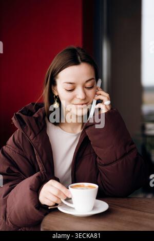 Young woman in a warm jacket talks on a smartphone while holding a cup of coffee at a cafe table. Casual lifestyle moment with soft light, indoor back Stock Photo