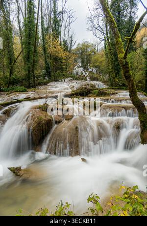 Long exposure photo of a cascading waterfall and river flowing over mossy rock terraces in a forest near Lods, France, with silky water, autumn trees Stock Photo