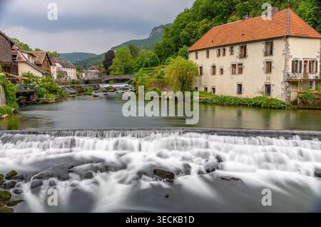 Long exposure view of a river and small waterfall flowing through Lods, France, with stone houses, bridge, lush green hills and calm water under an ov Stock Photo