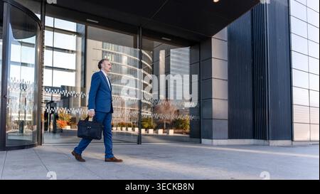 A middle-aged businessman in a blue suit walks confidently outside a modern office building, carrying a briefcase, with glass and steel architecture i Stock Photo