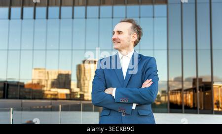 A confident middle-aged businessman in a blue suit stands with arms crossed, reflecting on modern urban architecture. The glass facade mirrors a vibra Stock Photo