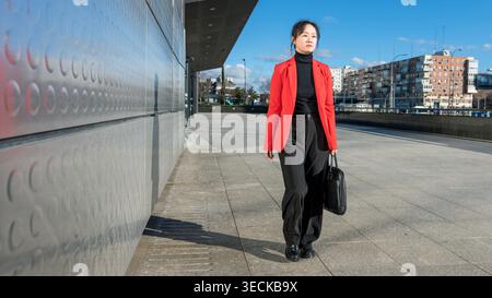Middle-aged businesswoman in a red blazer walking confidently along a modern city street. She exudes professionalism and determination, holding a blac Stock Photo